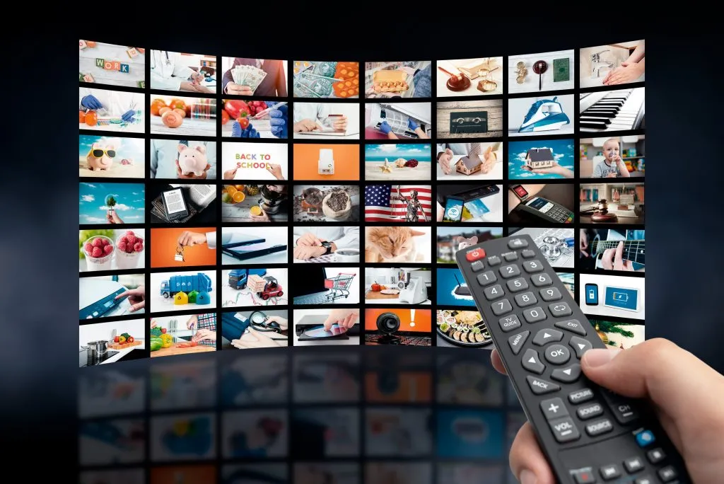 Hand hält Fernbedienung vor einer Wand aus TV-Bildschirmen mit verschiedenen Bildern.