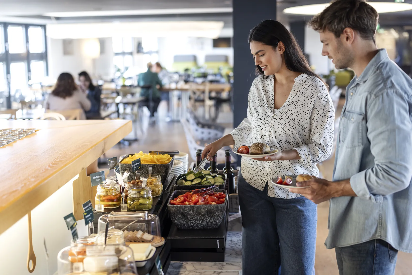 Two hotel guests serve themselves at the breakfast buffet with fresh vegetables and bread.