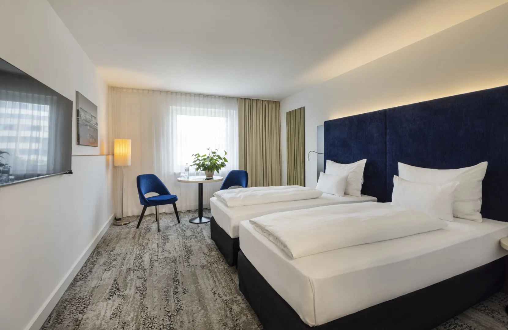 Bright hotel room with two single beds, blue chairs, table, and large window.